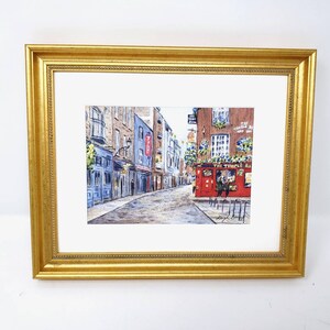May include: Watercolour painting of a European street scene, framed in a gold ornate frame. The artwork depicts a cobbled street lined with buildings, including a red pub with the text "The Temple Bar".