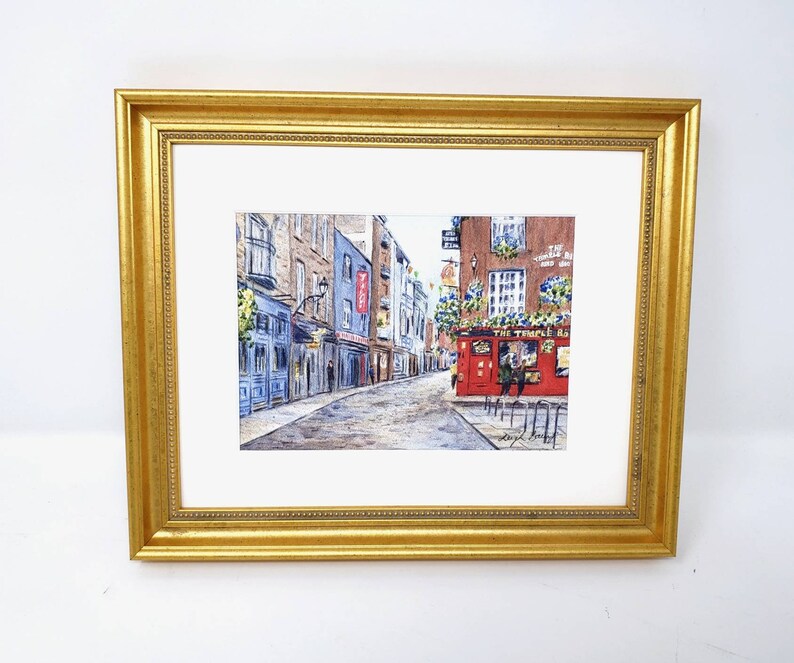 May include: Watercolour painting of a European street scene, framed in a gold ornate frame. The artwork depicts a cobbled street lined with buildings, including a red pub with the text "The Temple Bar".