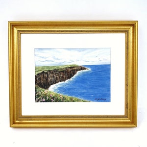 May include: Watercolor painting of a coastal landscape featuring a cliffside with green grass and brown rock formations. The ocean is a vibrant blue, and the artwork is framed in a gold-colored frame.