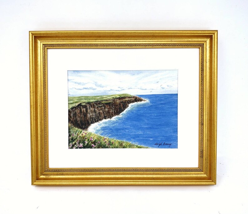 May include: Watercolor painting of a coastal landscape featuring a cliffside with green grass and brown rock formations. The ocean is a vibrant blue, and the artwork is framed in a gold-colored frame.