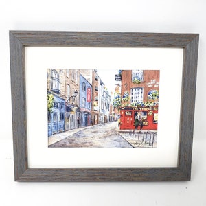 May include: Framed watercolour painting of a street scene. The artwork shows a cobblestone street with buildings in blue, brown, and red. A red pub with the text "The Temple Bar" is visible. The frame is made of gray wood.