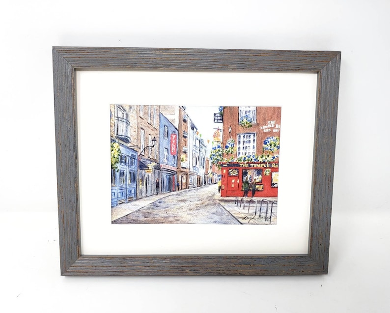May include: Framed watercolour painting of a street scene. The artwork shows a cobblestone street with buildings in blue, brown, and red. A red pub with the text "The Temple Bar" is visible. The frame is made of gray wood.