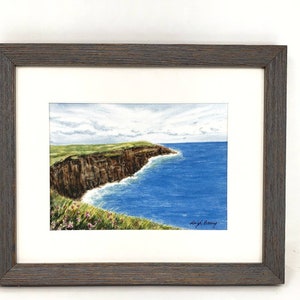 May include: A framed watercolor painting of a coastal landscape. The artwork depicts a brown cliffside meeting the blue ocean under a cloudy sky. Green grass and pink flowers are in the foreground. The frame is dark brown.