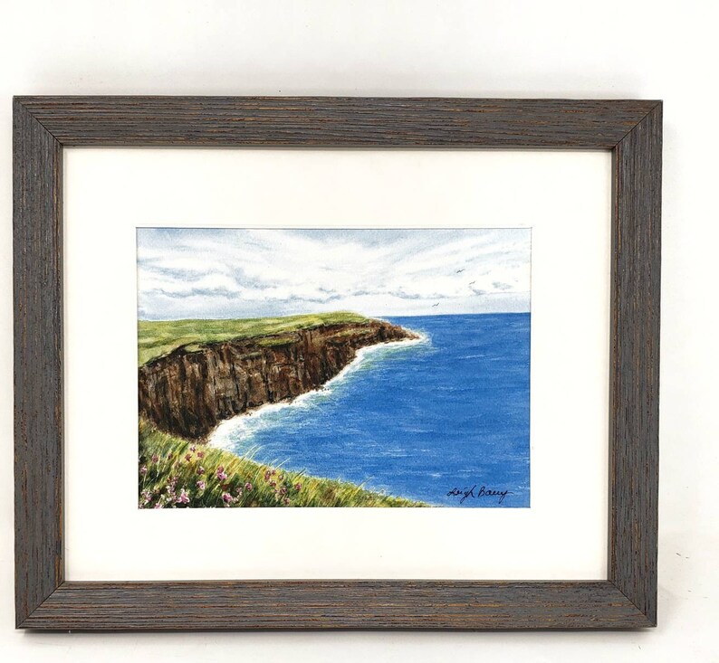 May include: A framed watercolor painting of a coastal landscape. The artwork depicts a brown cliffside meeting the blue ocean under a cloudy sky. Green grass and pink flowers are in the foreground. The frame is dark brown.