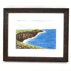 May include: Watercolor painting of a coastal landscape, framed in a dark brown wooden frame. The artwork depicts a cliffside with green grass, brown rock, and blue ocean. The sky is light blue with a few clouds. Signed by Leigh Barry.