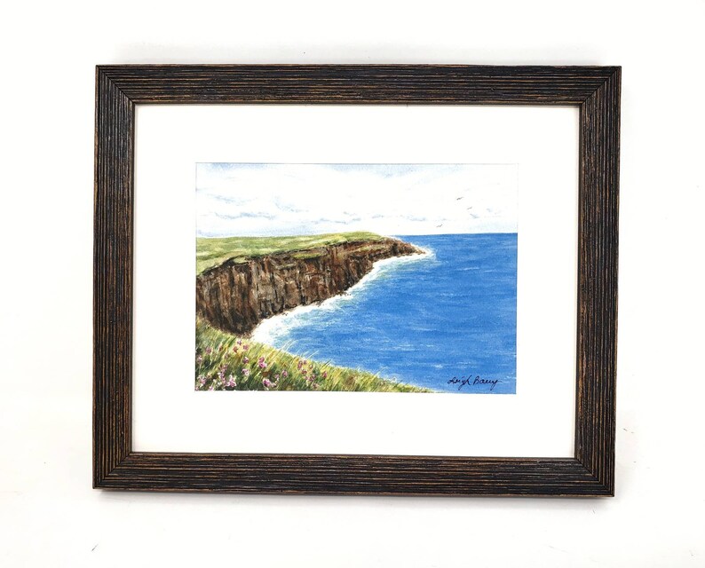 May include: Watercolor painting of a coastal landscape, framed in a dark brown wooden frame. The artwork depicts a cliffside with green grass, brown rock, and blue ocean. The sky is light blue with a few clouds. Signed by Leigh Barry.