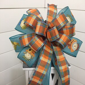 May include: A teal and orange plaid ribbon bow with a scarecrow pattern. The bow is tied around a white lantern.