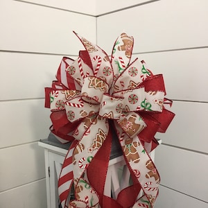 May include: A red and white striped ribbon with a gingerbread man pattern is tied into a bow. The bow is placed on top of a white lantern.