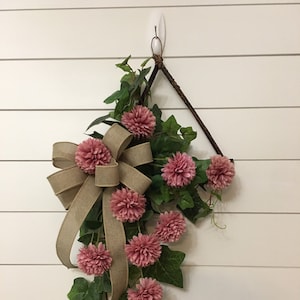 May include: A pink flower wreath with a burlap bow hanging on a white wall. The wreath is made of artificial flowers and greenery.