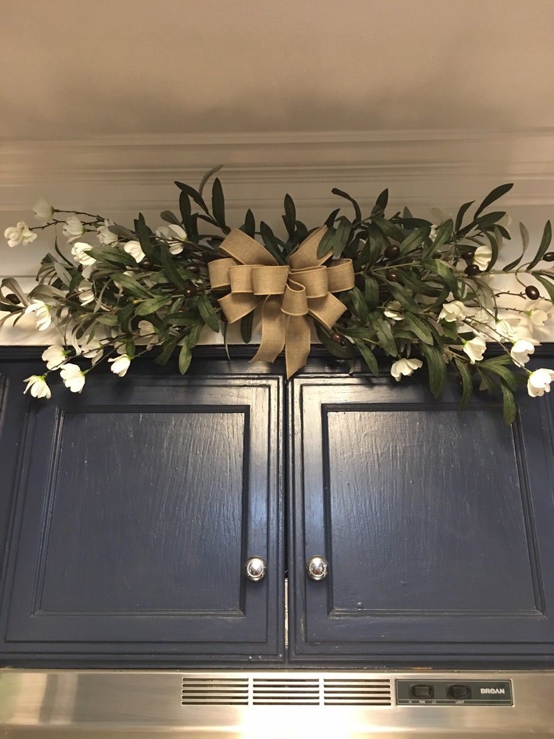May include: A kitchen cabinet with a blue painted finish and a garland of olive branches and white flowers with a burlap bow.