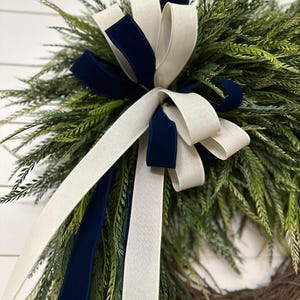 May include: A decorative wreath featuring green foliage and a large bow. The bow is made of white and navy blue velvet ribbon. The wreath is made of natural twigs and is suitable for indoor or outdoor use.