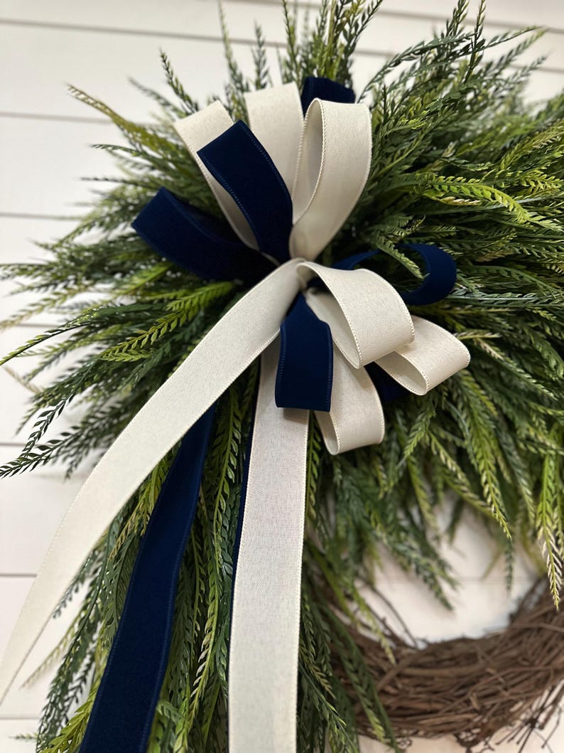 May include: A decorative wreath featuring green foliage and a large bow. The bow is made of white and navy blue velvet ribbon. The wreath is made of natural twigs and is suitable for indoor or outdoor use.