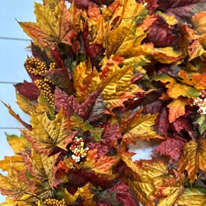 May include: A fall-themed wreath composed of colorful artificial leaves in shades of red, orange, and yellow. The wreath is adorned with small, round, golden-colored berries and white berries, creating a festive autumn decoration.