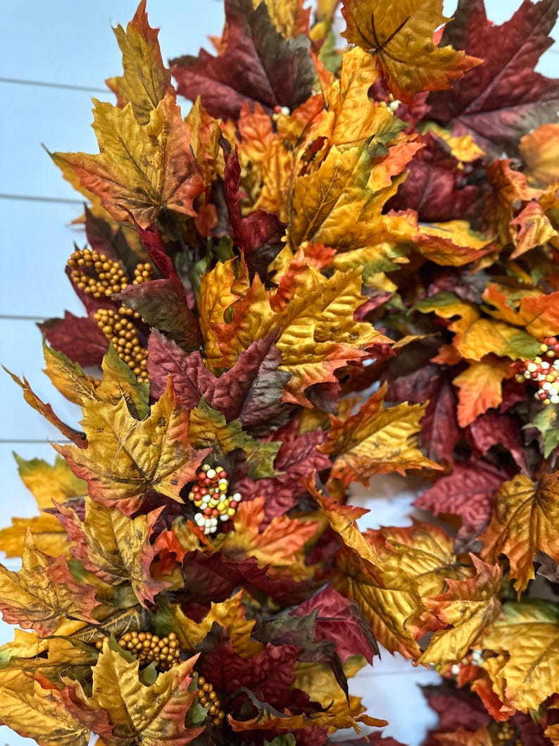 May include: A fall-themed wreath composed of colorful artificial leaves in shades of red, orange, and yellow. The wreath is adorned with small, round, golden-colored berries and white berries, creating a festive autumn decoration.