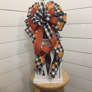 May include: A black and white buffalo plaid ribbon with orange ribbon and orange and yellow fall leaves printed ribbon tied into a bow. The bow is on top of a white lantern.