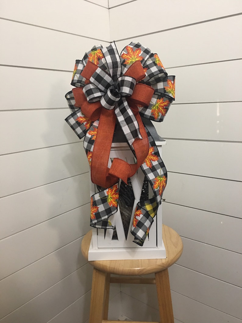 May include: A black and white buffalo plaid ribbon with orange ribbon and orange and yellow fall leaves printed ribbon tied into a bow. The bow is on top of a white lantern.