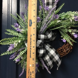 May include: A black and white checkered ribbon tied around a wreath of lavender sprigs. The wreath is 14 inches in diameter. The wreath is hanging on a dark blue door.