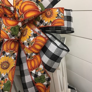 May include: A black and white buffalo plaid ribbon bow with a pumpkin and sunflower print ribbon. The bow is tied around a white lantern.