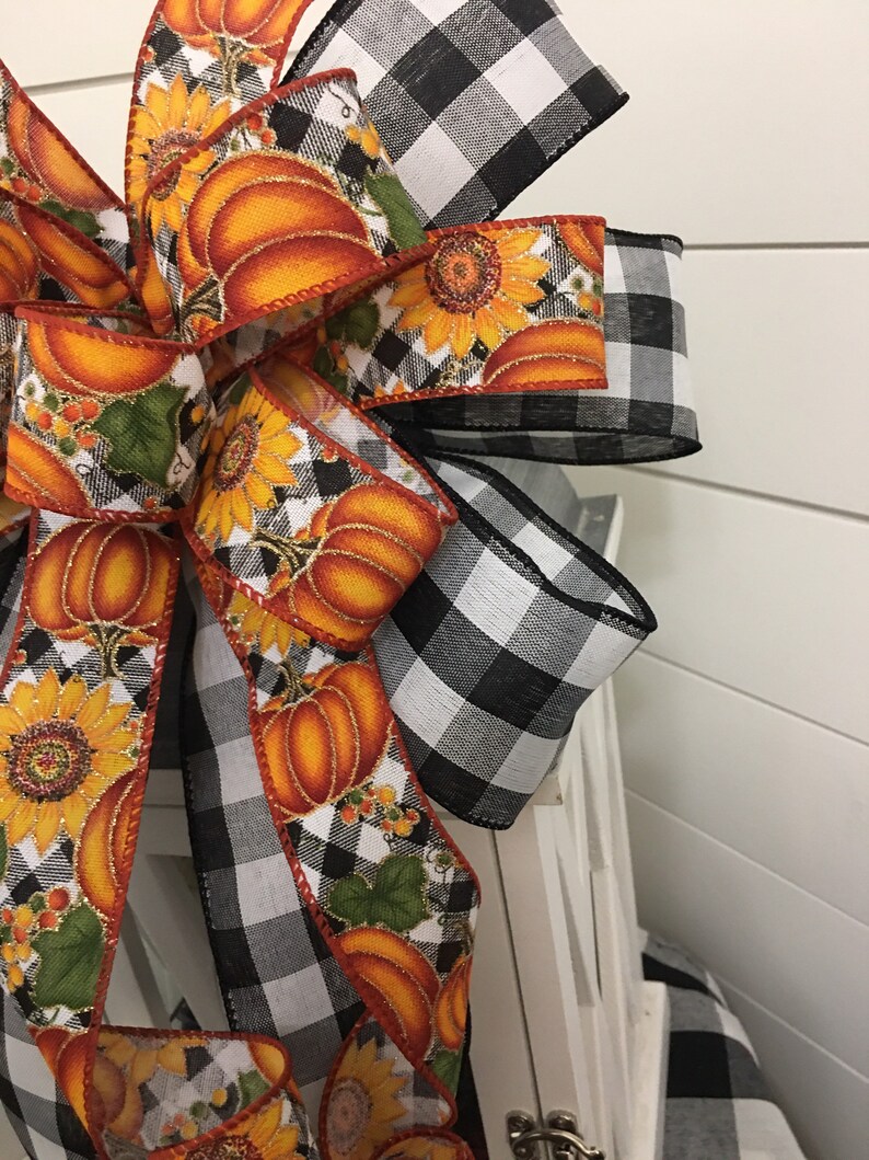 May include: A black and white buffalo plaid ribbon bow with a pumpkin and sunflower print ribbon. The bow is tied around a white lantern.