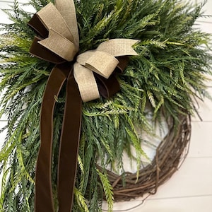 May include: A decorative wreath featuring a natural grapevine base, adorned with lush green faux foliage. A large bow made of tan and brown ribbons adds a touch of elegance. Perfect for seasonal home decor.