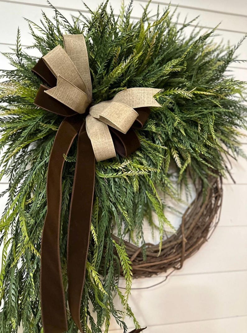 May include: A decorative wreath featuring a natural grapevine base, adorned with lush green faux foliage. A large bow made of tan and brown ribbons adds a touch of elegance. Perfect for seasonal home decor.