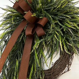 May include: A decorative wreath featuring a woven grapevine base, lush green foliage, and a rich brown ribbon bow. The wreath's design incorporates varying shades of green and brown, creating a natural and inviting aesthetic. Perfect for seasonal home decor.