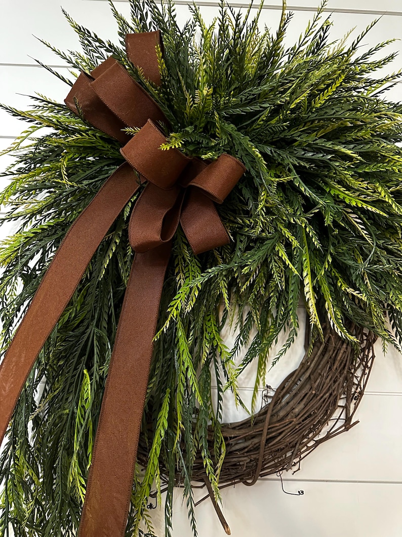 May include: A decorative wreath featuring a woven grapevine base, lush green foliage, and a rich brown ribbon bow. The wreath's design incorporates varying shades of green and brown, creating a natural and inviting aesthetic. Perfect for seasonal home decor.