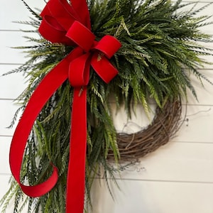 May include: A decorative wreath featuring a base of interwoven brown twigs, adorned with lush green foliage. A vibrant red velvet ribbon is tied in a large bow at the top and cascades down the side, adding a festive touch. The wreath is displayed against a white background.