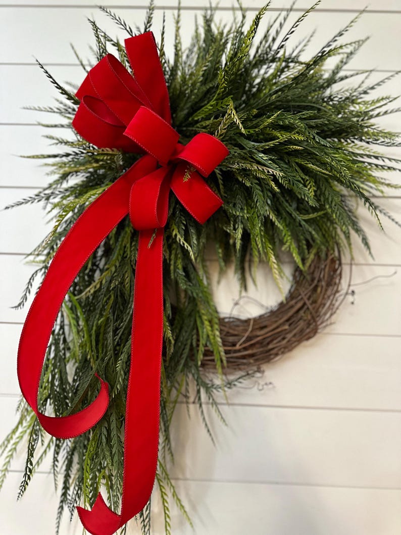 May include: A decorative wreath featuring a base of interwoven brown twigs, adorned with lush green foliage. A vibrant red velvet ribbon is tied in a large bow at the top and cascades down the side, adding a festive touch. The wreath is displayed against a white background.