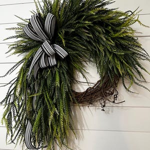 May include: A decorative wreath featuring lush green foliage and a prominent black and white striped bow. The wreath is constructed on a natural grapevine base, creating a rustic and inviting aesthetic. Perfect for seasonal home decor.