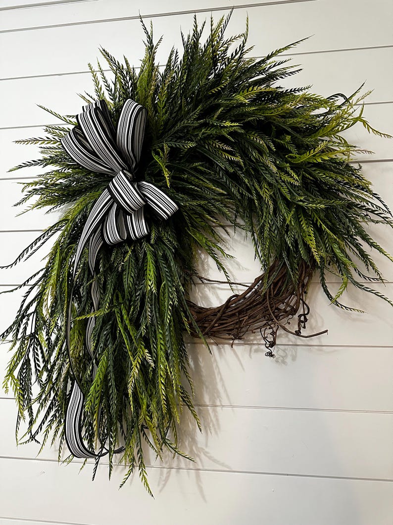 May include: A decorative wreath featuring lush green foliage and a prominent black and white striped bow. The wreath is constructed on a natural grapevine base, creating a rustic and inviting aesthetic. Perfect for seasonal home decor.
