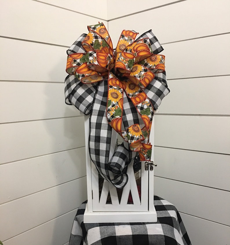 May include: A white lantern with a black and white checkered bow with a pumpkin and sunflower print. The bow is tied around the top of the lantern.