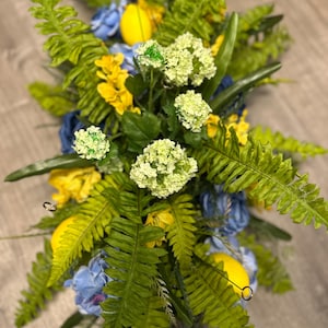 May include: A vibrant floral arrangement featuring a mix of artificial flowers and greenery. The arrangement includes yellow, blue, and white flowers, along with green fern-like leaves and yellow lemon-shaped accents. The design is symmetrical and suitable for home decor.
