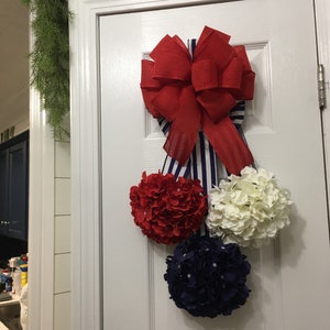 May include: A patriotic door decoration featuring a large red bow with blue and white striped ribbon. Below the bow are three floral spheres in red, white, and navy blue, creating a festive and decorative display.