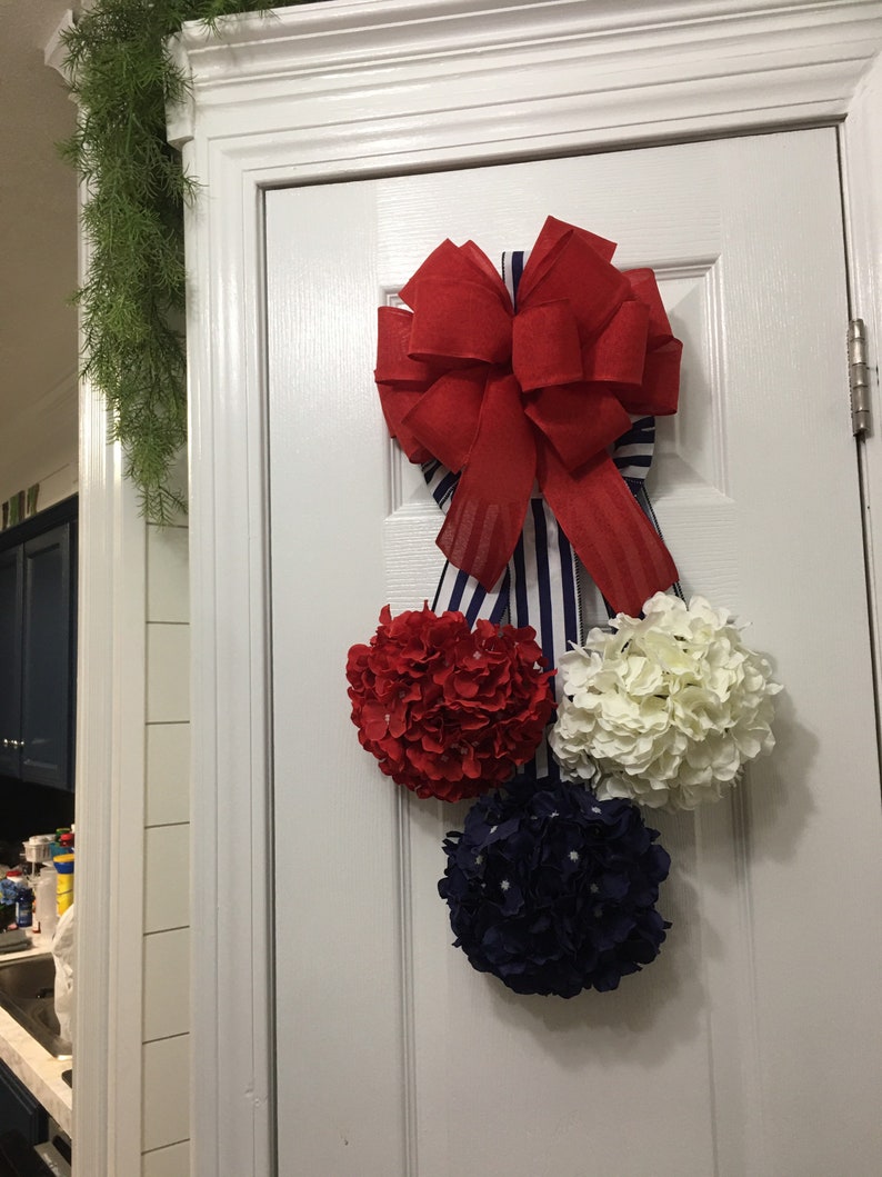 May include: A patriotic door decoration featuring a large red bow with blue and white striped ribbon. Below the bow are three floral spheres in red, white, and navy blue, creating a festive and decorative display.