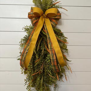 May include: A fall-themed decorative swag with a large mustard yellow bow and plaid ribbon accents. The swag features green foliage, orange berries, and dried grasses, creating a rustic and seasonal aesthetic. The background is a white wooden wall.