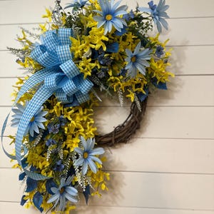 May include: A spring wreath featuring yellow and blue flowers, accented with a blue and white gingham ribbon. The wreath is made of woven twigs and is displayed against a white wooden background.