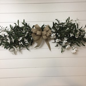 May include: A white wall with a green and white floral garland with a brown burlap bow.