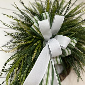 May include: A close-up of a decorative wreath featuring green and white striped ribbon and a large white bow. The wreath is composed of various shades of green foliage, creating a natural and inviting aesthetic. The white background provides a clean contrast.