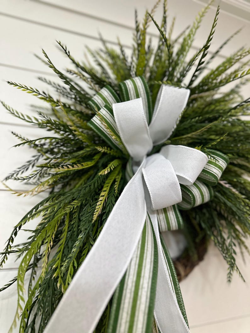 May include: A close-up of a decorative wreath featuring green and white striped ribbon and a large white bow. The wreath is composed of various shades of green foliage, creating a natural and inviting aesthetic. The white background provides a clean contrast.