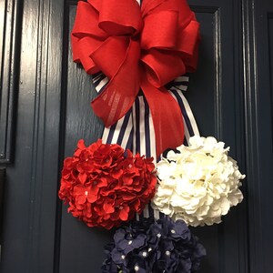 May include: A patriotic-themed door decoration featuring a large red bow with a striped ribbon. Below the bow are three clusters of artificial hydrangeas in red, white, and navy blue, creating a festive display.