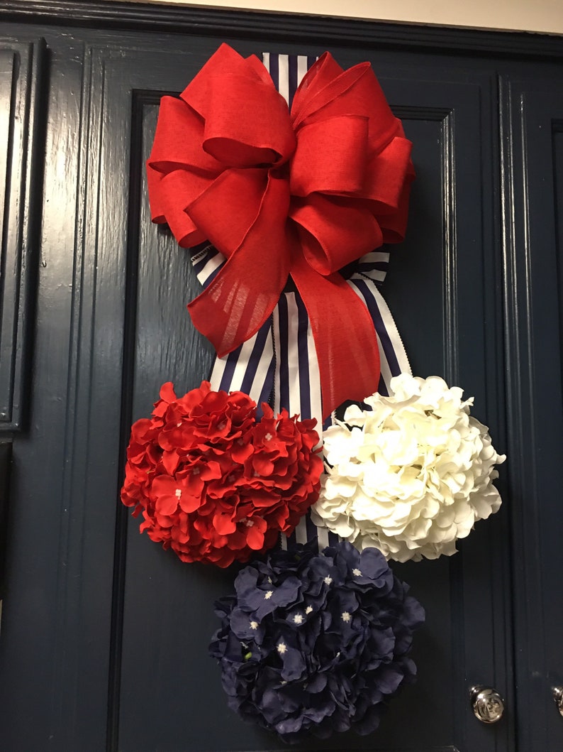 May include: A patriotic-themed door decoration featuring a large red bow with a striped ribbon. Below the bow are three clusters of artificial hydrangeas in red, white, and navy blue, creating a festive display.