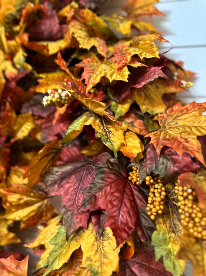 May include: Close-up of a fall-themed garland with maple leaves in shades of red, orange, and yellow. The garland also features small, round, yellow berries. The leaves have detailed veining and a realistic appearance.