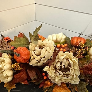 May include: A fall-themed floral arrangement featuring cream-colored peonies, orange pumpkins, and vibrant autumn leaves. The arrangement includes berries and wheat stalks, creating a warm, seasonal display.