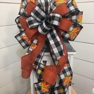 May include: A black and white buffalo plaid ribbon bow with orange and yellow fall leaves printed on a white ribbon. The bow is tied around a white lantern.