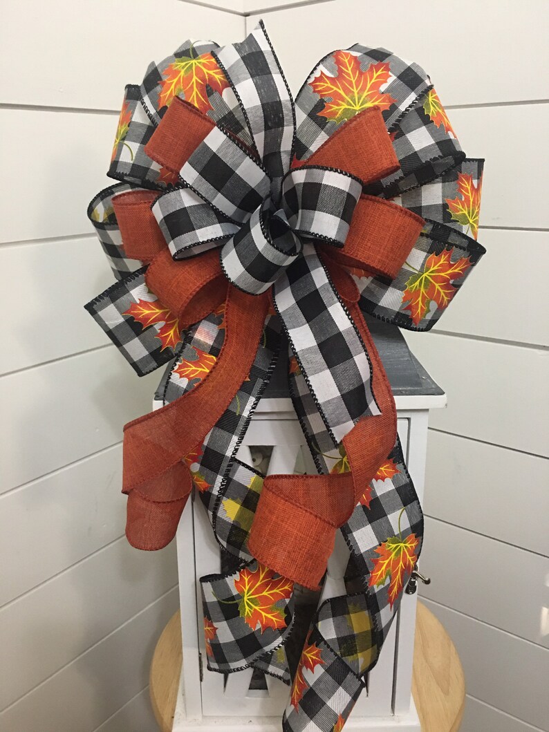 May include: A black and white buffalo plaid ribbon bow with orange and yellow fall leaves printed on a white ribbon. The bow is tied around a white lantern.