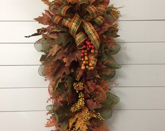 May include: A fall-themed swag decoration featuring faux leaves in shades of brown, orange, and green, accented with berries and a plaid ribbon bow. The swag is designed to hang vertically, creating a festive autumn display.