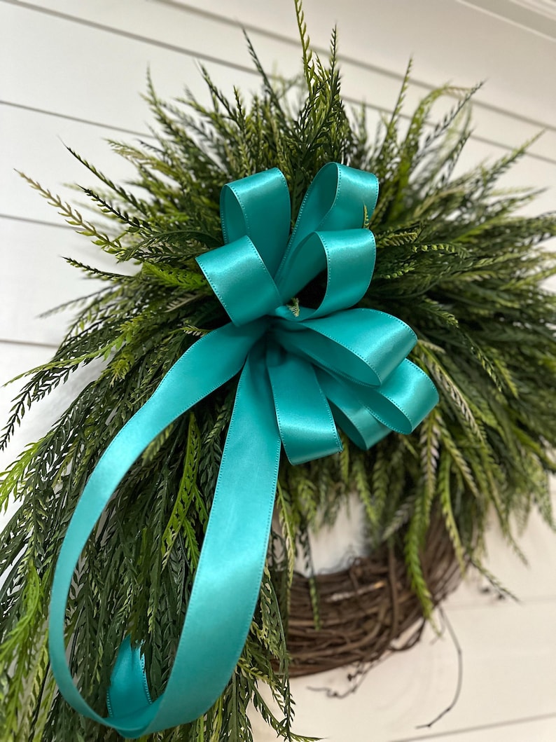 May include: A decorative wreath featuring a teal satin bow and lush green foliage. The wreath is made of woven brown branches, creating a natural and inviting aesthetic. Perfect for adding a touch of elegance to any home decor.