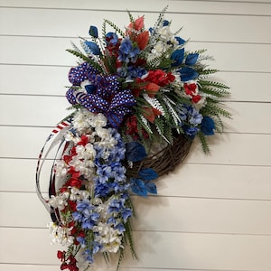 May include: A patriotic wreath with a brown grapevine base, adorned with red, white, and blue faux flowers and greenery. A large bow with a star pattern and ribbon accents the design. The wreath is designed to hang on a door or wall.
