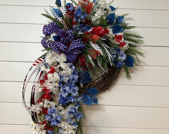 May include: A patriotic wreath with a brown grapevine base, adorned with red, white, and blue faux flowers and greenery. A large bow with a star pattern and ribbon accents the design. The wreath is designed to hang on a door or wall.
